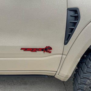 Pode incluir: Um close-up de um caminhão bege com um logotipo "TRX" vermelho e uma ventilação lateral preta. O caminhão tem um pneu preto com um padrão de banda de rodagem.