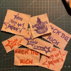 Puede incluir: Un conjunto de pequeños parches de tela beige con bordes irregulares. Cada parche presenta texto escrito a mano en naranja y morado, incluyendo frases como "FACK THE RICH" y "FROS SUR LA PATATE", junto con diseños de estrellas. Los parches están sobre una esterilla de corte verde.