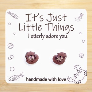May include: A white card with the text "It's Just Little Things I otterly adore you" and a pair of brown otter earrings. The card also has a small crab and fish illustrations.