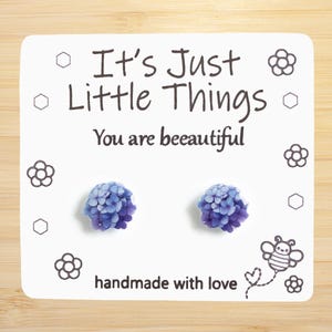 May include: A pair of purple flower stud earrings with the text "It's Just Little Things You are bee-autiful" and "handmade with love" on a white card.