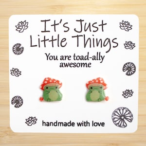 May include: A pair of green frog earrings with red and white polka dot mushroom hats. The earrings are handmade and come with a card that says "It's Just Little Things You are toad-ally awesome handmade with love."