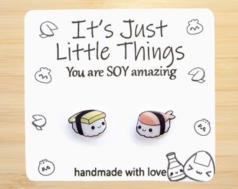 May include: A white card with black text that reads "It's Just Little Things You are SOY amazing" and a pair of handmade sushi earrings. The earrings are made of clear plastic and feature a cute cartoon face. One earring is a yellow and white maki roll and the other is a pink and white nigiri with a shrimp on top.