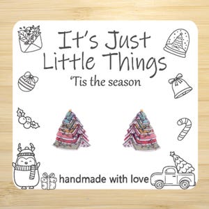 May include: A white card with the text "It's Just Little Things" and "'Tis the season". The card features illustrations of Christmas ornaments, a penguin, and two Christmas tree earrings. The words "handmade with love" are also present.