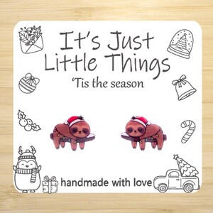 May include: A white card with the text "It's Just Little Things" and "'Tis the season". The card features illustrations of Christmas-themed items, including a penguin, a snow globe, and two sloths wearing Santa hats. The words "handmade with love" are also present.