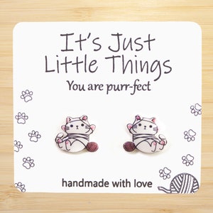 May include: A pair of white cat-shaped earrings with pink accents. The earrings are on a white card with the text "It's Just Little Things You are purr-fect" and "handmade with love".