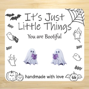 May include: A pair of white ghost earrings with purple books, on a white card with the text "It's Just Little Things You are Bootiful" and a black and white illustration of a spider web, ghosts, and pumpkins.