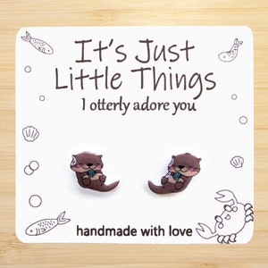Op de afbeelding: Oorbelletjes in de vorm van een otter op een witte kaart met de tekst "It's Just Little Things I otterly adore you" en "handmade with love". De oorbellen zijn bruin met roze accenten en houden een blauwe schelp vast.