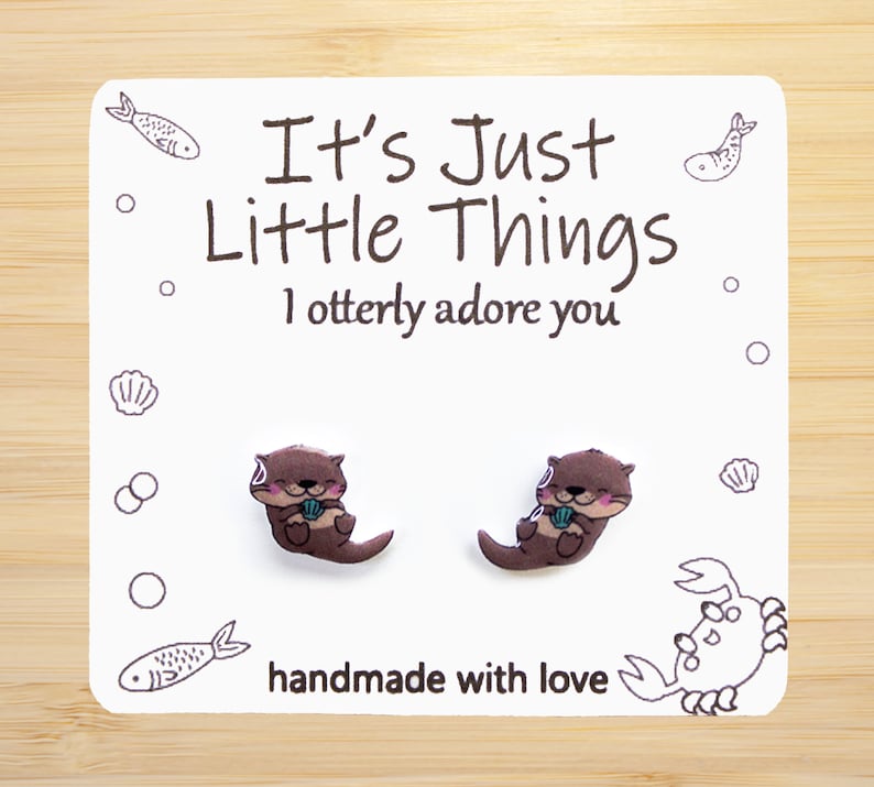 Op de afbeelding: Oorbelletjes in de vorm van een otter op een witte kaart met de tekst "It's Just Little Things I otterly adore you" en "handmade with love". De oorbellen zijn bruin met roze accenten en houden een blauwe schelp vast.
