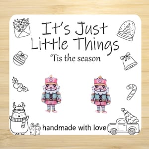 May include: A white card with the text "It's Just Little Things" and "'Tis the season". The card features illustrations of Christmas ornaments and two nutcracker earrings. The phrase "handmade with love" is also present.