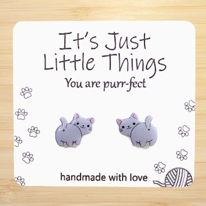 May include: A white card with the text "It's Just Little Things You are purr-fect" and two gray cat butt earrings. The card also has paw prints and a ball of yarn.