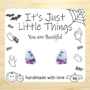May include: A pair of handmade earrings featuring a ghost reading a book. The earrings are made with polymer clay and have a pink and white color scheme. The card in the background says "It's Just Little Things You are Bootiful" and is decorated with ghosts and bats.