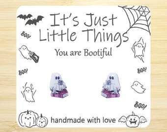 May include: A pair of handmade earrings featuring a ghost reading a book. The earrings are made with polymer clay and have a pink and white color scheme. The card in the background says "It's Just Little Things You are Bootiful" and is decorated with ghosts and bats.