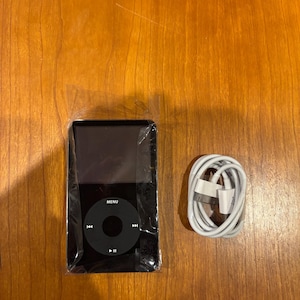Puede incluir: iPod negro con un panel de control circular y una pantalla, empaquetado en plástico. Un cable de carga blanco está enrollado al lado. El dispositivo tiene un botón "MENU" y controles de reproducción.