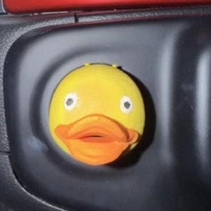 May include: A yellow duck-shaped object with an orange beak and white eyes. The duck is set against a dark background, possibly a car's interior. The object appears to be a decorative item or accessory.