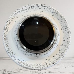May include: A Nest smart thermostat with a black screen and a silver bezel, set in a white, speckled ceramic base. The base has a circular shape with a wide rim, featuring a terrazzo-like pattern of gray and white chips.