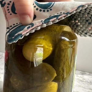 Könnte beinhalten: Ein großes Glas mit grünen Essiggurken. Ein dekorativer Stoff-Gläseröffner mit einem rosa- und türkisblauen Paisley-Muster wird über den Deckel des Glases gelegt. Die Gurken sind in einer klaren Salzlake getaucht.
