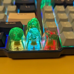 May include: A close-up of a keyboard with four keycaps replaced with clear, colorful keycaps. The keycaps are green, blue, and red, and they are illuminated from below. The keyboard is black and has a white and gray color scheme.