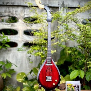 Puede incluir: Un instrumento musical tailandés tradicional, un Phin, con un cuerpo rojo y un clavijero de madera tallada. El instrumento está sostenido por un soporte negro, y una tarjeta con una ilustración es visible.