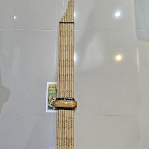 Könnte beinhalten: Ein hohes Musikinstrument aus Bambus mit mehreren zusammengebundenen Pfeifen. Das Instrument hat ein dunkles Mundstück aus Holz und dekorative Muster, die in den Bambus geätzt sind. Eine kleine Karte liegt in der Nähe des Sockels.