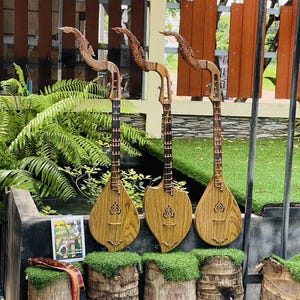 Puede incluir: Tres instrumentos de cuerda de madera con cabezas talladas en forma de dragón. Los instrumentos tienen un acabado marrón claro con detalles en marrón oscuro. Se exhiben al aire libre, apoyados en una repisa de piedra y tocones de madera.