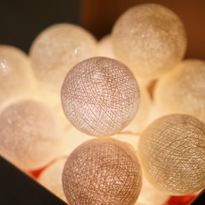 Puede incluir: Un conjunto de luces de cadena esféricas brillantes en una caja oscura. Las luces son una mezcla de blanco y beige, con una textura tejida. Ideal para la decoración del hogar.