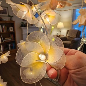May include: A decorative string of flower lights. Each flower features a delicate, layered petal design in white and yellow, with a small light bulb at the centre. The lights are strung on a thin, light-coloured wire, creating a warm, inviting glow.