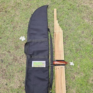 Puede incluir: Un instrumento tradicional de flauta de pan de bambú con una funda de transporte negra. El instrumento está hecho de múltiples tubos de bambú unidos. Una pieza de madera está unida a los tubos de bambú.