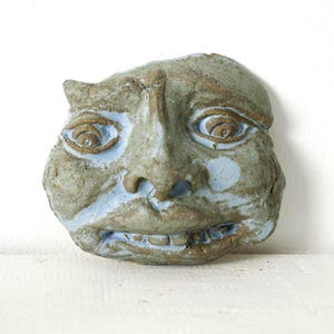 Puede incluir: Una escultura de rostro de cerámica con un aspecto desgastado y texturizado. La cara es principalmente gris verdoso con acentos azules alrededor de los ojos, la nariz y la boca. La escultura tiene la boca ligeramente abierta y un aspecto caprichoso.
