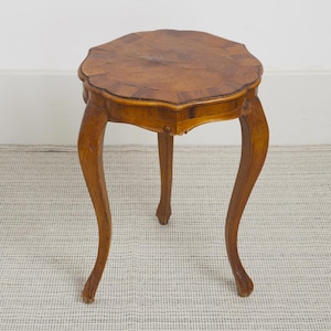 Op de afbeelding: Een kleine houten bijzettafel met een rond, geschulpt tafelblad met een zichtbaar houtnerfpatroon. De tafel wordt ondersteund door drie slanke, gebogen poten die vanuit het midden naar buiten uitlopen en een stabiele basis vormen.