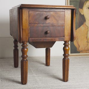 Puede incluir: Una mesa auxiliar de madera marrón con dos cajones apilados y pequeños pomos redondos. La mesa tiene cuatro patas torneadas y laterales abatibles que amplían la superficie. La veta de la madera es visible en toda la pieza con un acabado pulido.
