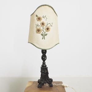 Puede incluir: Lámpara de mesa vintage con una pantalla de color crema con flores amarillas bordadas y hojas verdes. La lámpara tiene una base oscura y ornamentada y un tallo retorcido. Iluminación decorativa.