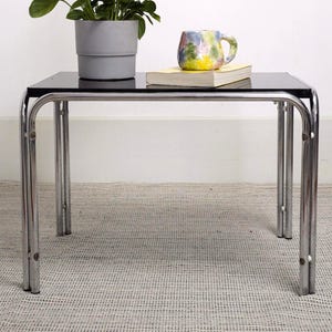 May include: A modern black glass coffee table with a chrome frame. A potted green plant and a colorful mug rest on the table. The mug sits on top of a yellow book. The table is on a light-colored rug.