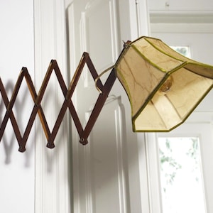 Peut inclure: Une lampe accordéon murale dotée d'un mécanisme à ciseaux en bois brun foncé qui s'étend vers l'extérieur. La lampe comprend un abat-jour en tissu beige à facettes multiples avec une bordure verte et une ampoule visible à l'intérieur.