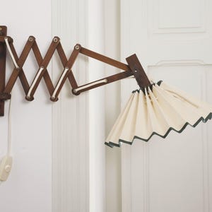 Peut inclure: Une lampe murale vintage en bois avec un bras extensible en accordéon. L'abat-jour plissé est de couleur crème avec une bordure vert foncé. Elle est fixée à un mur blanc, avec un cordon blanc et un interrupteur marche/arrêt.