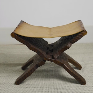 Può includere: Sgabello in legno fatto a mano con seduta in pelle marrone chiaro. Le gambe marrone scuro sono scolpite con un motivo strutturato. Lo sgabello è progettato con una base a forma di X per la stabilità.