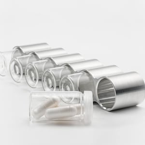 Puede incluir: Un vial de vidrio transparente que contiene dos cápsulas blancas, junto a una serie de soportes de metal plateado. Las cápsulas son cilíndricas con extremos redondeados. Los soportes tienen un diseño curvo.
