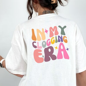 May include: A white short-sleeved t-shirt featuring a multi-colored graphic on the back with the text "IN MY CLOGGING ERA" in a bold, wavy font. The letters are colored in shades of orange, yellow, pink, purple, and green, accented by small stars.