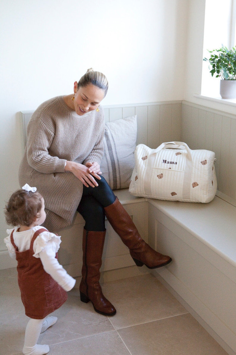 May include: A woman wearing a brown sweater and brown boots sits on a bench with a white diaper bag that says "Mama" on it. A young girl in a brown dress stands in front of the bench.