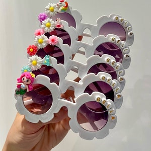 May include: Five pairs of white plastic sunglasses with purple lenses and a flower and other embellishments on the side. Each pair has the name "BELLA" spelled out in gold letters on the side.