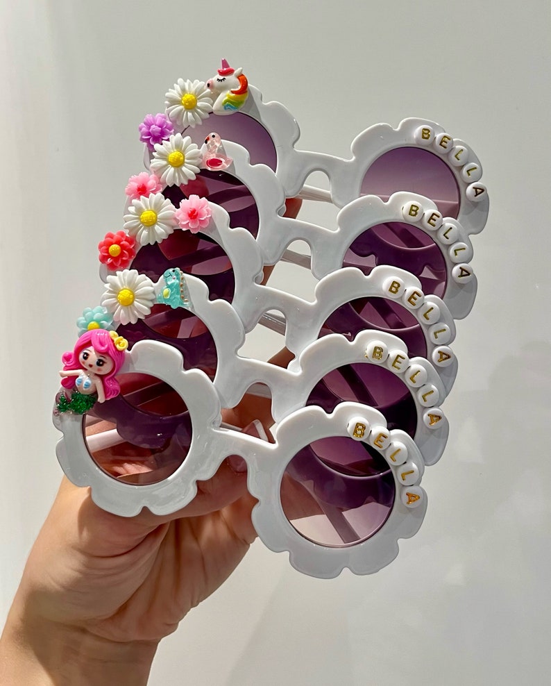 May include: Five pairs of white plastic sunglasses with purple lenses and a flower and other embellishments on the side. Each pair has the name "BELLA" spelled out in gold letters on the side.