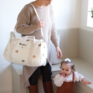 May include: A white diaper bag with brown bear print and the word "Mama" embroidered on the front. A woman in a brown sweater and brown boots is holding the bag. A young girl in a white shirt and brown overalls is standing next to her.