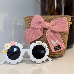 May include: White sunglasses with a floral design and black lenses. The sunglasses have the name "BELLA" spelled out in individual letter beads. A pink bow and a woven bag are also in the image.
