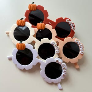Peut inclure: Plusieurs paires de lunettes de soleil en forme de fleur, de couleurs blanche, pêche et rouge. Chaque paire est ornée d'une petite citrouille orange et d'un prénom en perles blanches. Les verres sont noirs.