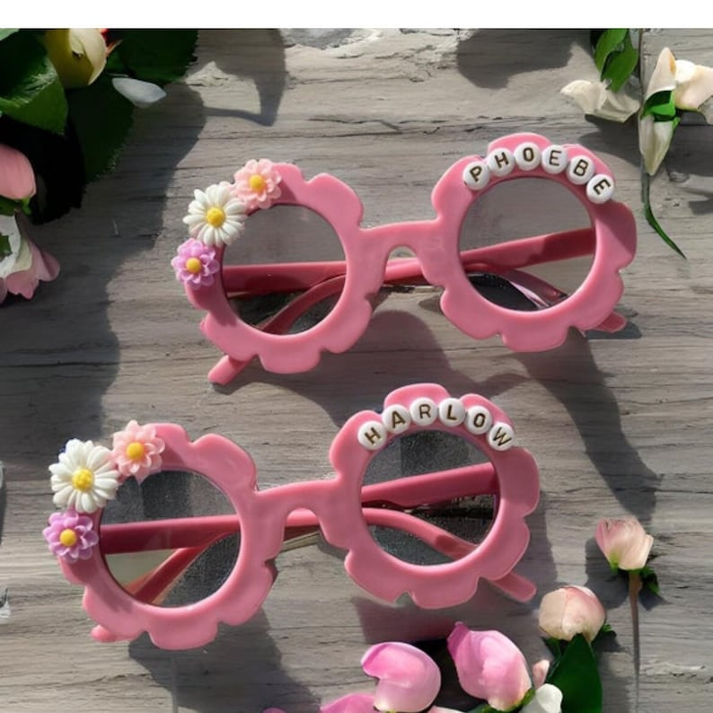 May include: Two pairs of pink plastic sunglasses with flower accents. The top pair says "PHOEBE" in gold letters and the bottom pair says "HARLOW" in gold letters.