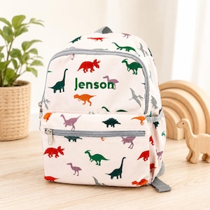 May include: A cream-coloured backpack featuring a repeating pattern of colourful dinosaurs in shades of red, green, orange, and purple. The front pocket displays the name Jenson in dark green text, with grey zippers and side pockets.