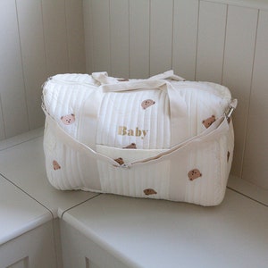 May include: A white quilted diaper bag with brown bear embroidery and the word "Baby" embroidered in gold. The bag has two shoulder straps and a zipper closure.