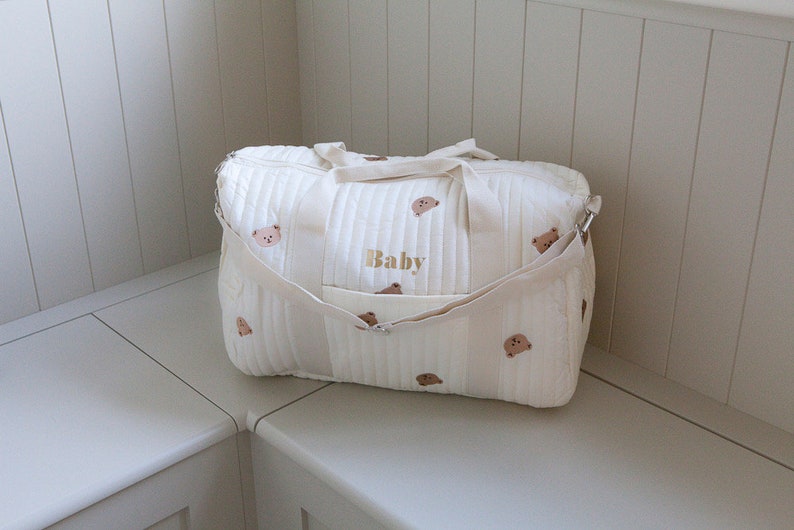 May include: A white quilted diaper bag with brown bear embroidery and the word "Baby" embroidered in gold. The bag has two shoulder straps and a zipper closure.
