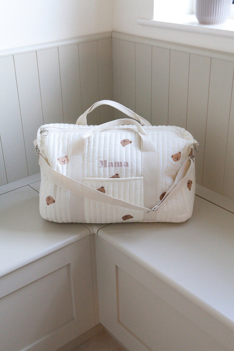 May include: A white quilted travel bag with brown bear illustrations and the word "Mama" embroidered in pink. The bag has a zippered pocket and a long strap.