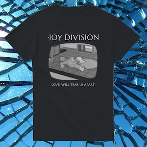 May include: Black t-shirt featuring the band name "JOY DIVISION" above a grayscale image of a person lying on a rug. The text "LOVE WILL TEAR US APART" is below the image. The shirt is made of a soft material.
