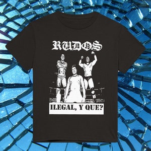 Peut inclure: T-shirt noir avec un motif blanc. Le motif présente le mot "RUDOS" au-dessus d'une scène de lutte et le texte "ILEGAL, Y QUE?" en dessous. L'image représente trois lutteurs sur un ring.